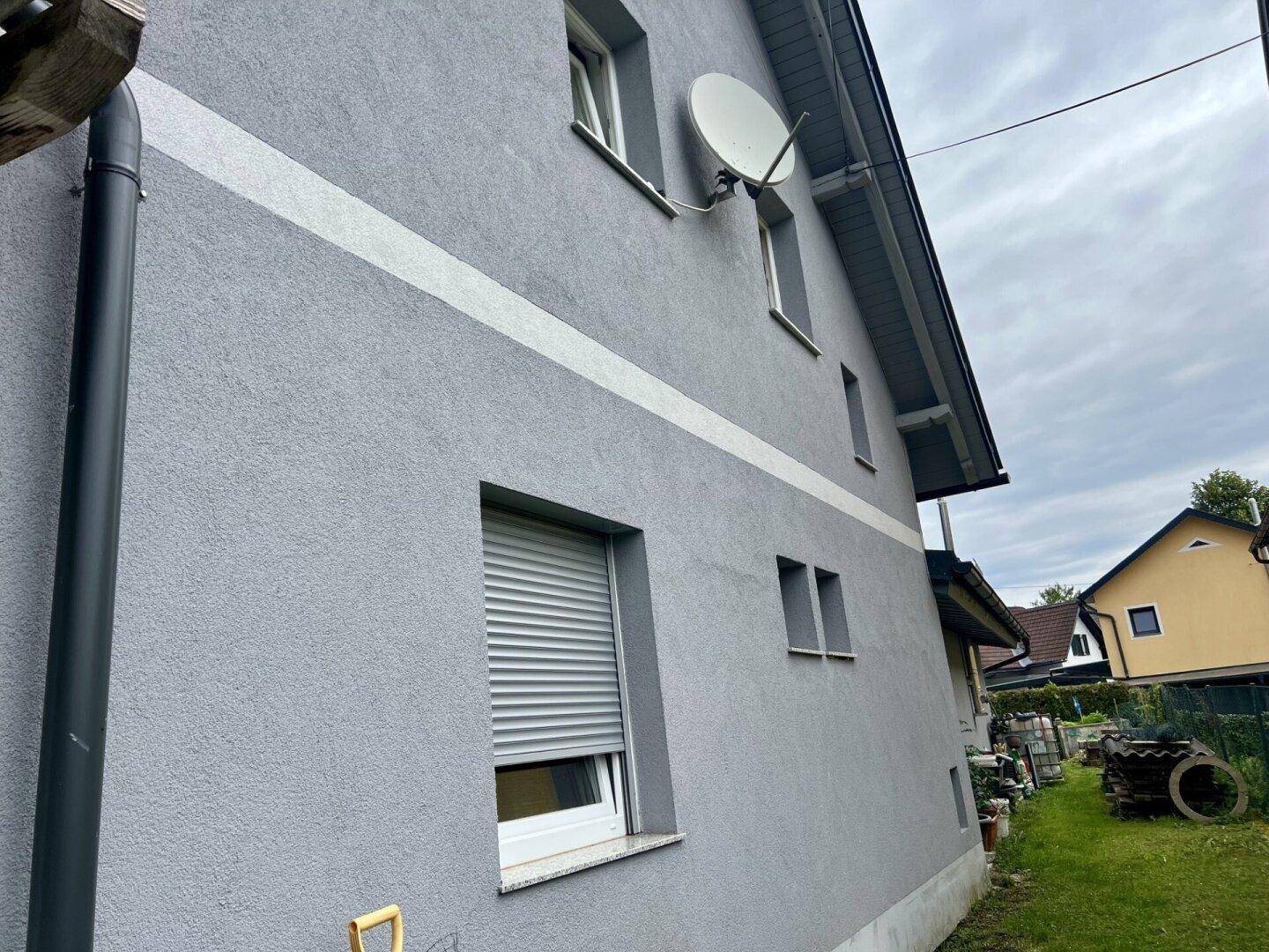 Seitenansicht des Hauses mit grauer Fassade, Fenstern und einem Satellitenempfänger.