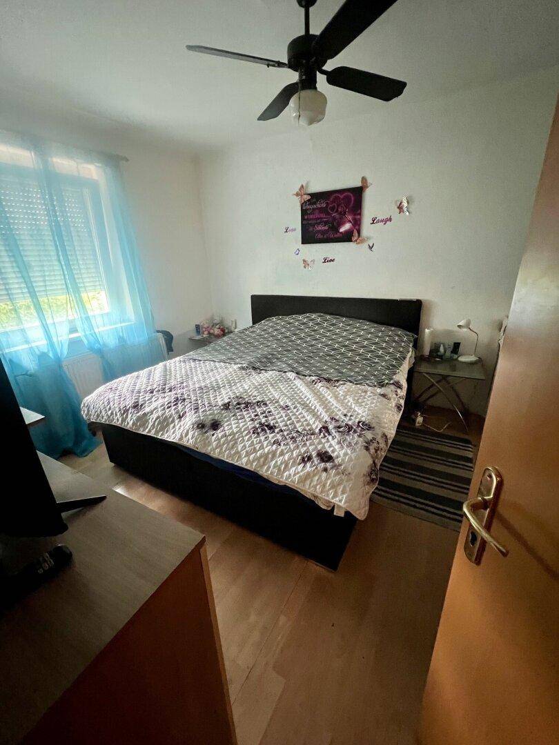 Helles Schlafzimmer mit Doppelbett, Laminatboden und einem Fenster mit blauen Vorhängen.