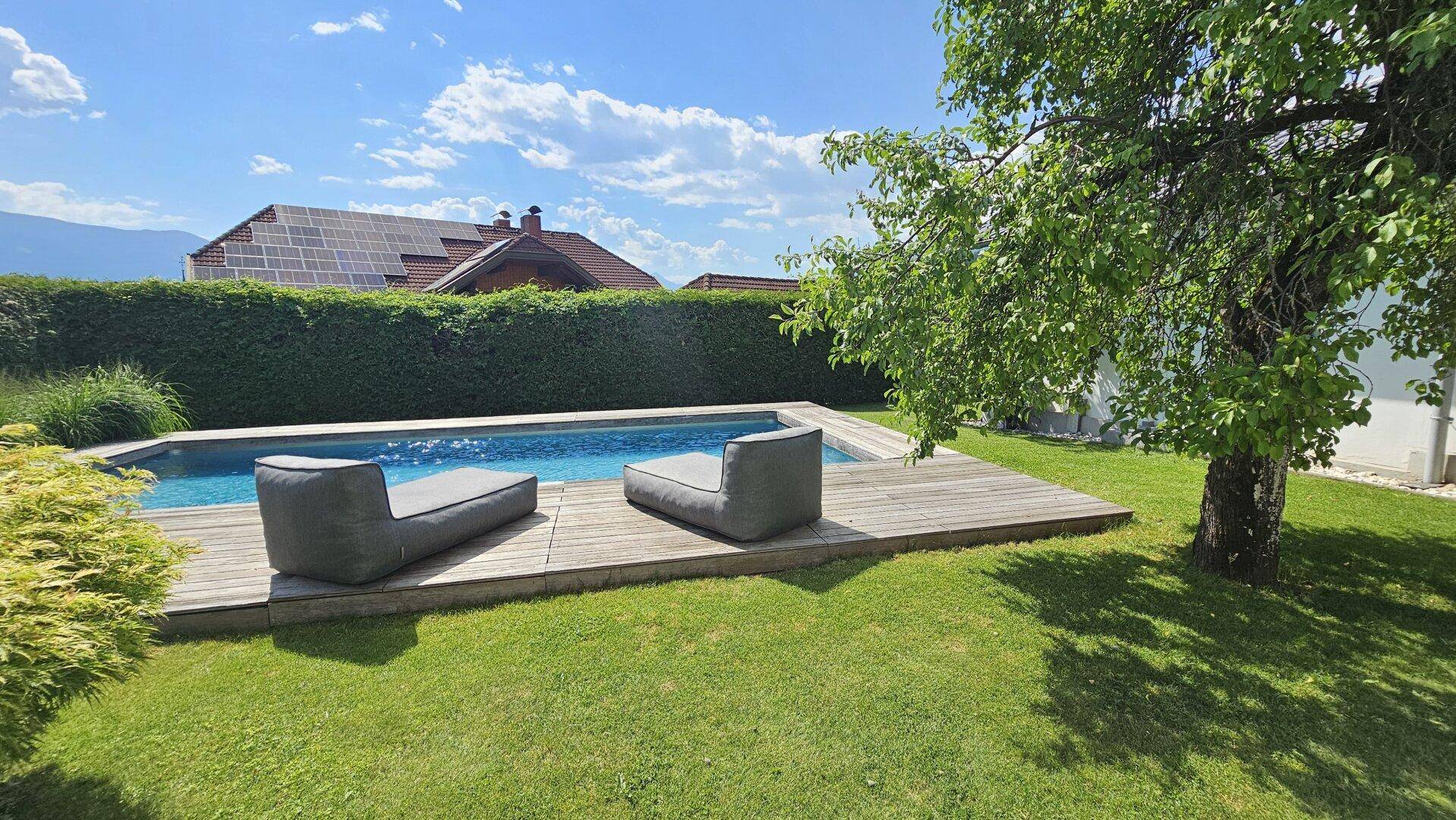 Entspannender Poolbereich mit Holzdeck und Liegen, umgeben von grünem Rasen und hohen Hecken.
