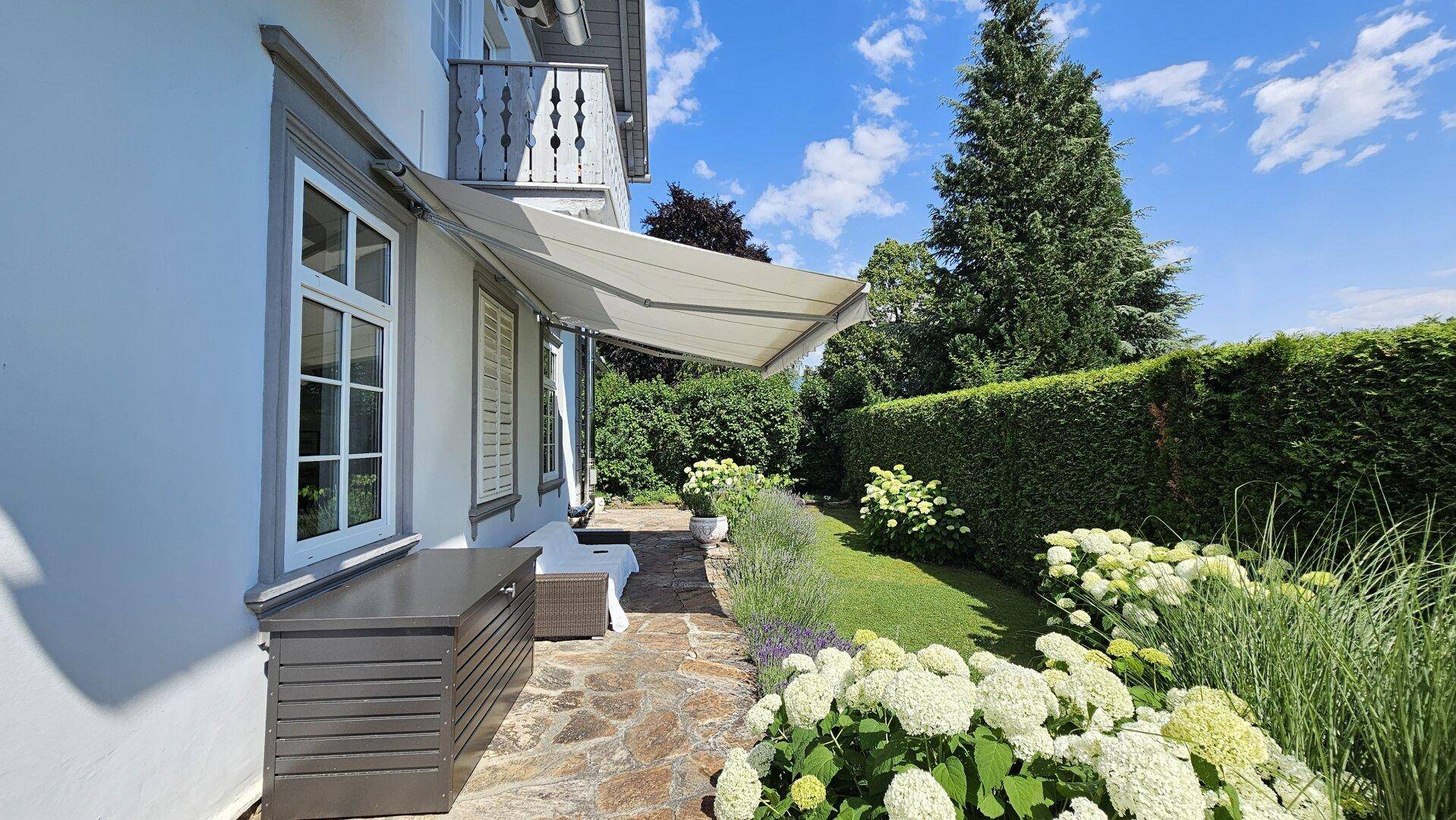 Seitlicher Gartenbereich mit Steinweg, ausfahrbarer Markise und blühenden Hortensien unter blauem Himmel.