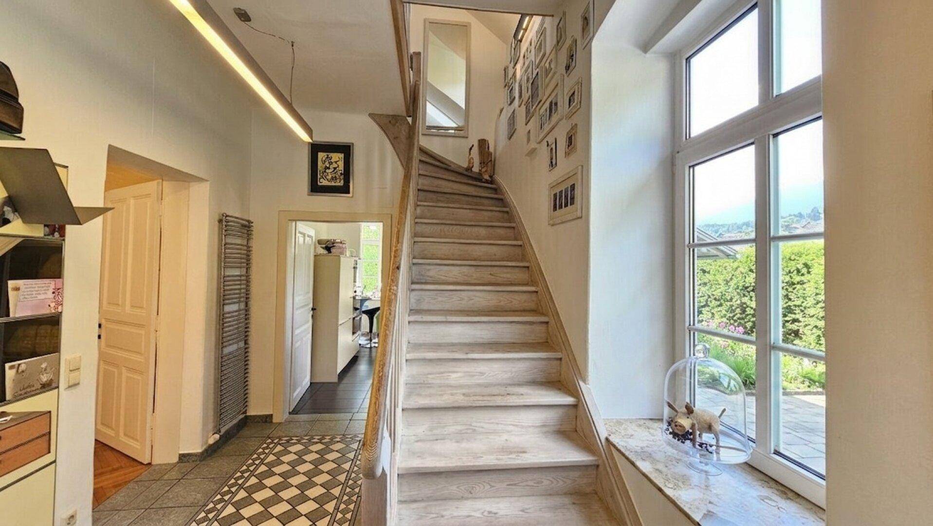 Heller Flur mit eleganter Holztreppe, gemustertem Fliesenboden und großem Fenster für viel Tageslicht.