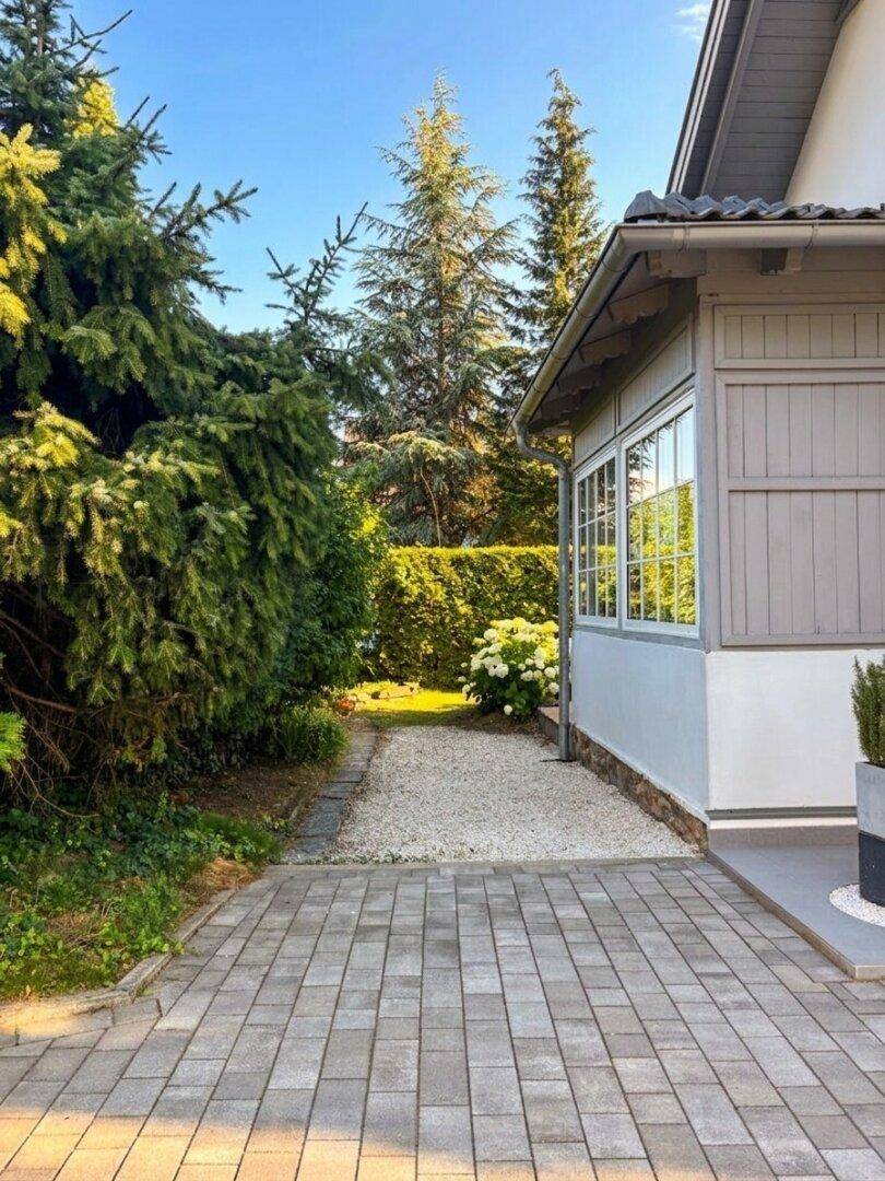 Gepflegter Kiesweg neben dem Haus, umgeben von üppigem Grün und hohen Bäumen.