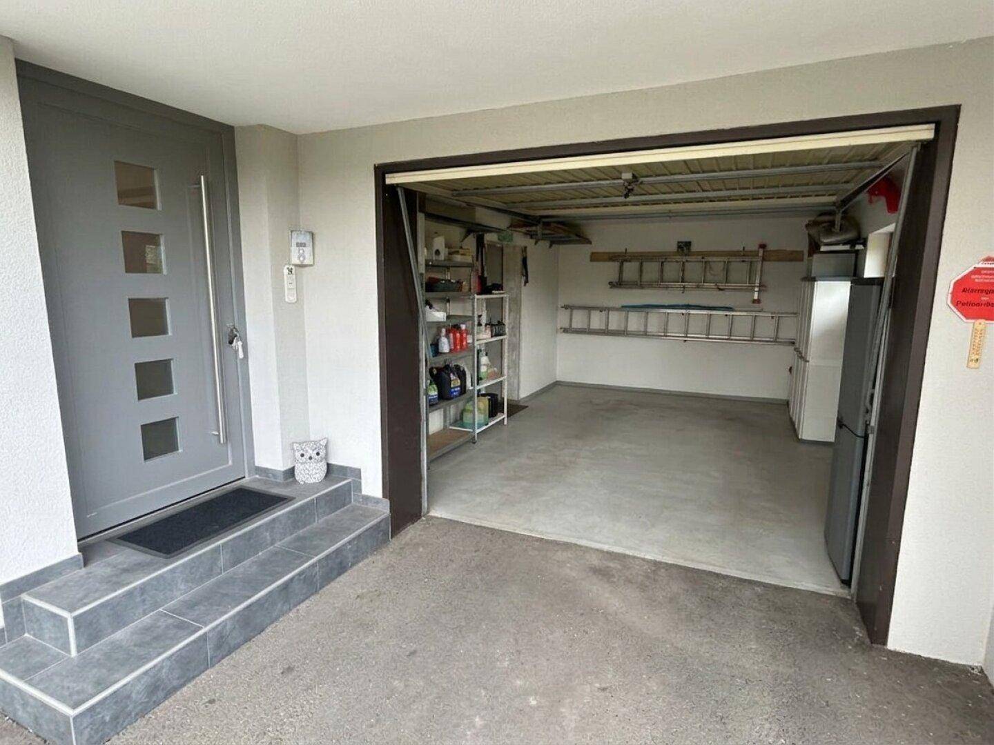 Eingangsbereich mit moderner grauer Tür und offener Garage mit Stauraum.