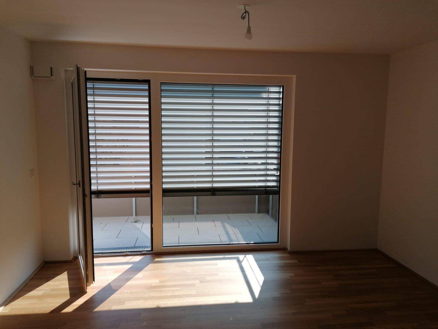Heller Raum mit Holzboden und großer Fensterfront, die Zugang zu einem Außenbereich bietet.