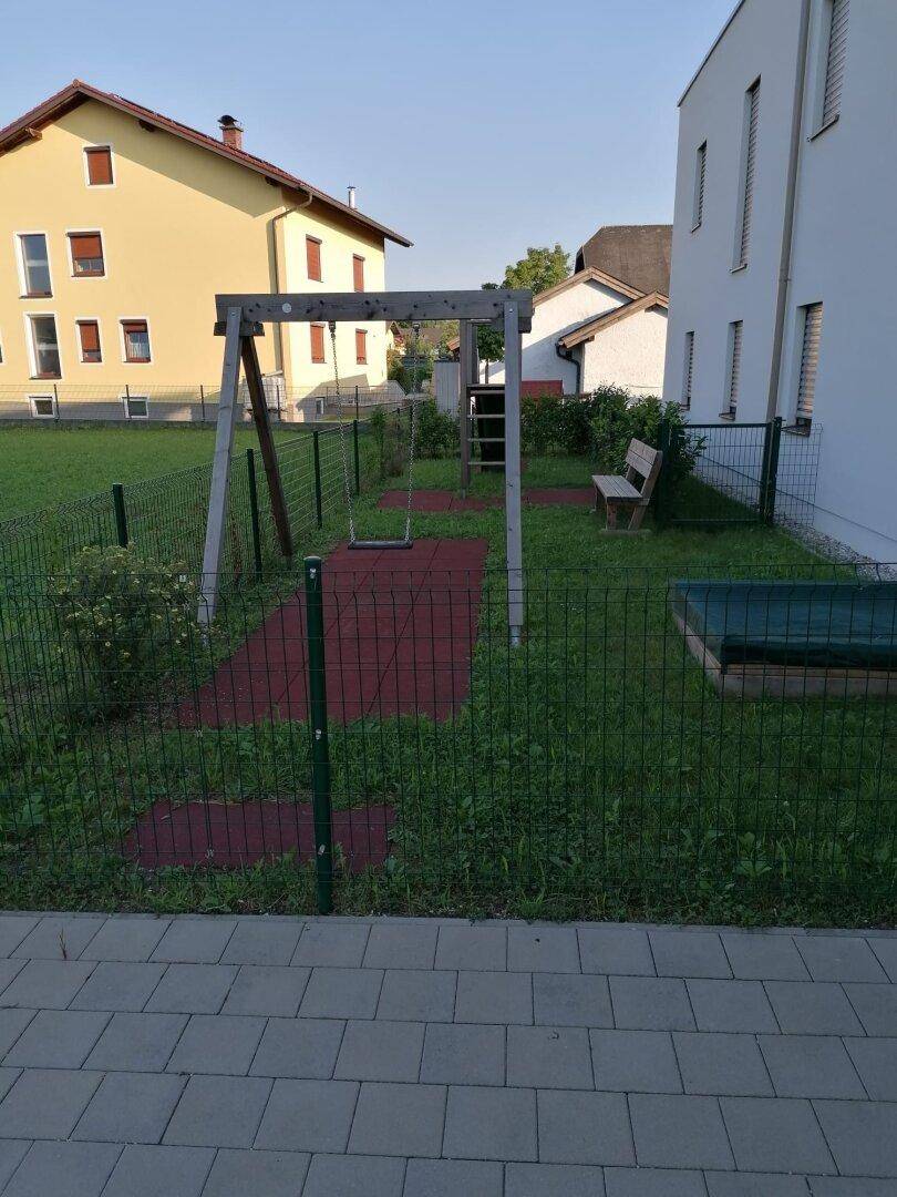 Kleiner Spielplatz mit Schaukel und Sandkasten, umgeben von Rasen und einem Zaun.
