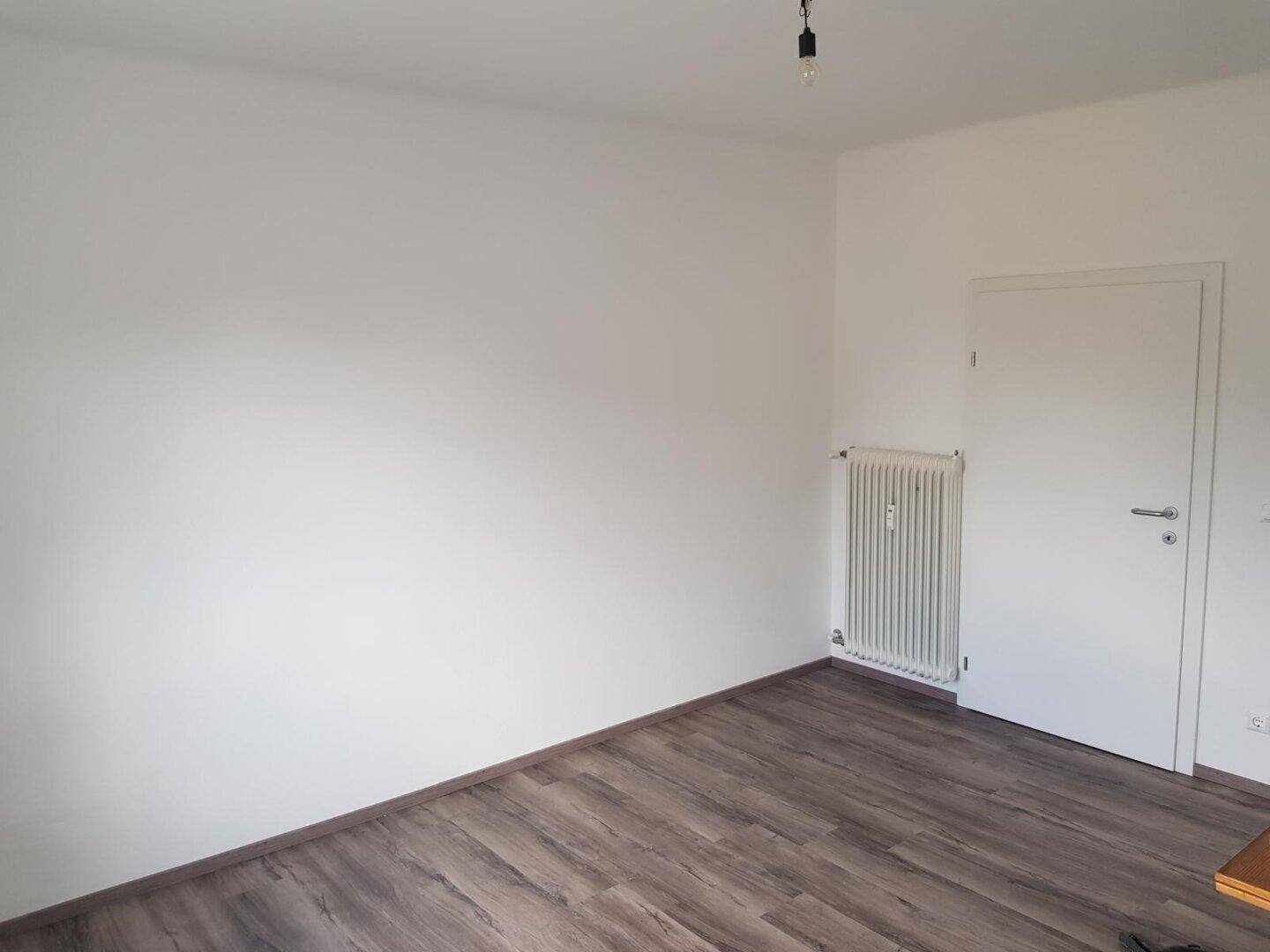 Helles, leeres Zimmer mit dunklem Laminatboden und Heizkörper.