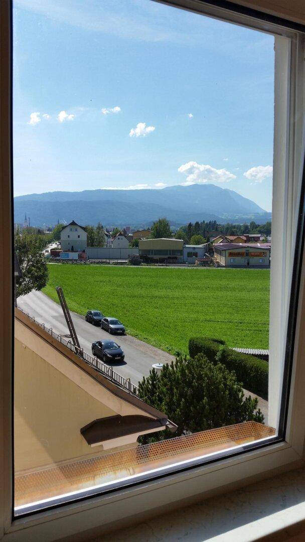 Blick aus dem Fenster auf eine grüne Wiese, eine Straße und Berge in der Ferne.