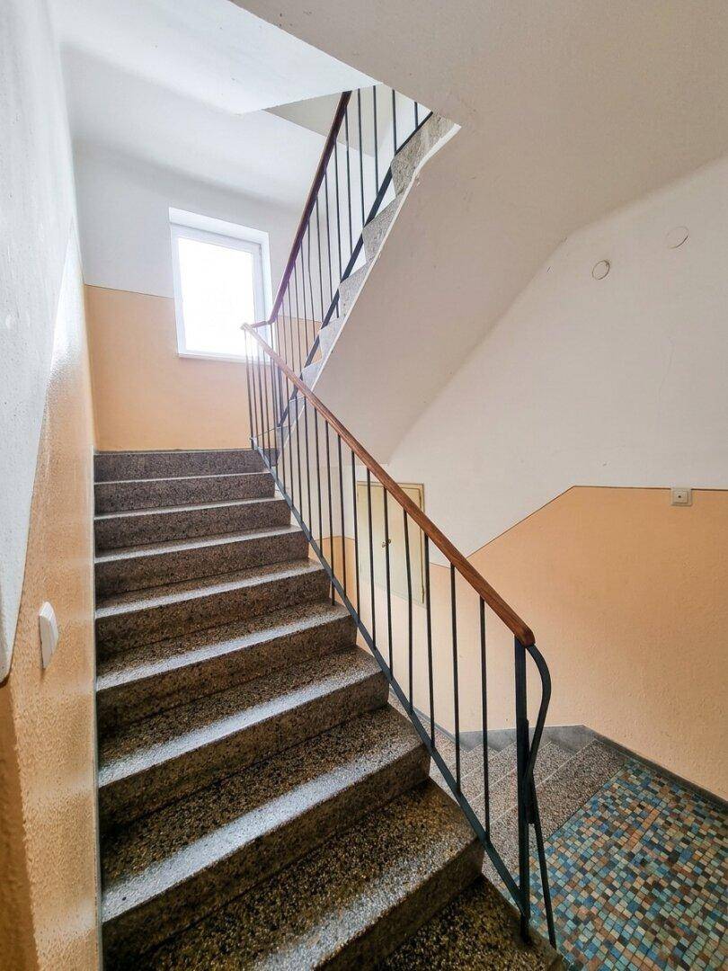 Treppenhaus mit Terrazzostufen, Metallgeländer und einem Fenster, das Tageslicht hereinlässt.