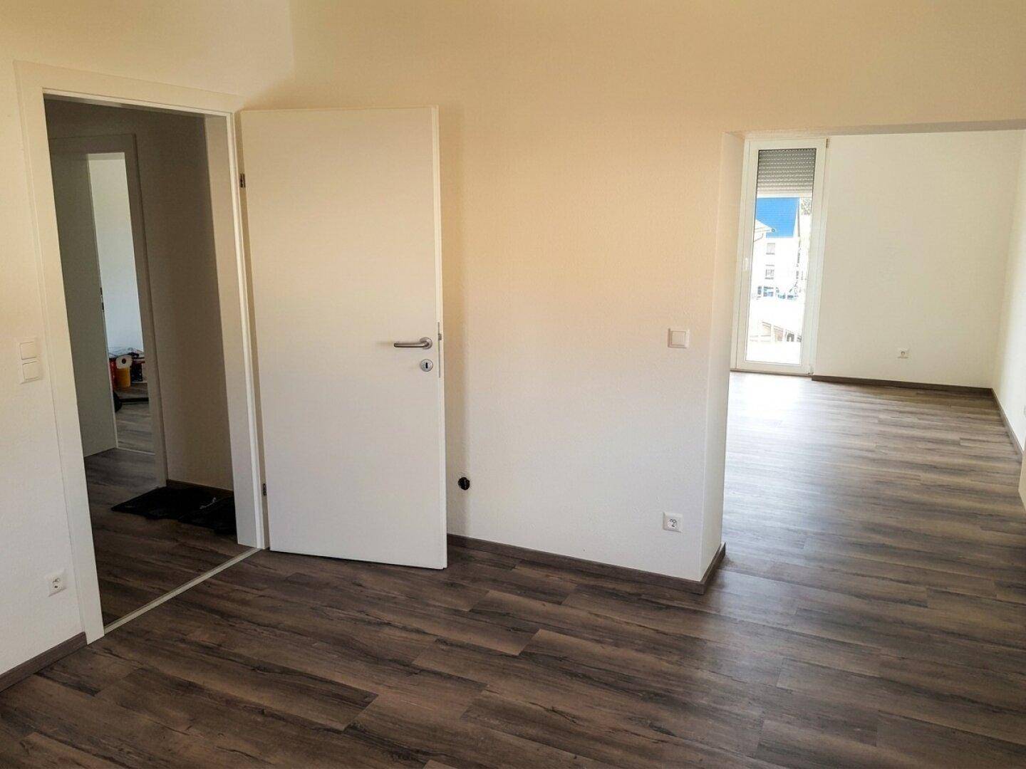 Geräumiges Zimmer mit dunklem Laminatboden und Zugang zu einem weiteren Raum.