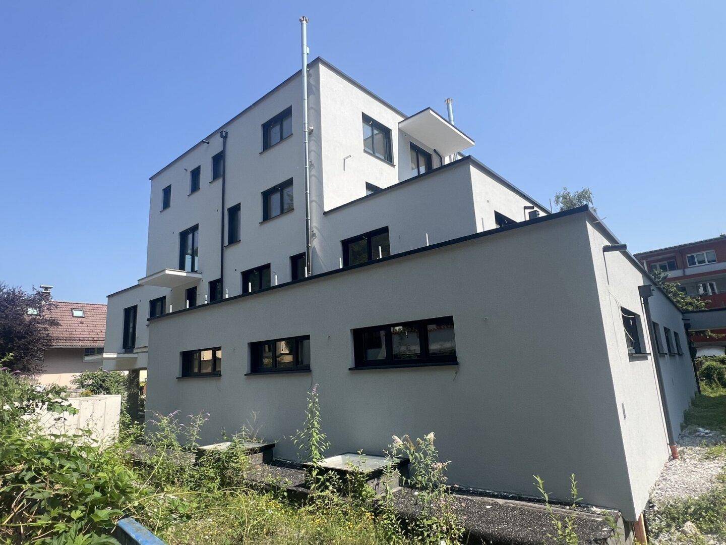 Seitenansicht eines modernen Neubaus mit grauer Fassade, Flachdächern und Balkonen.