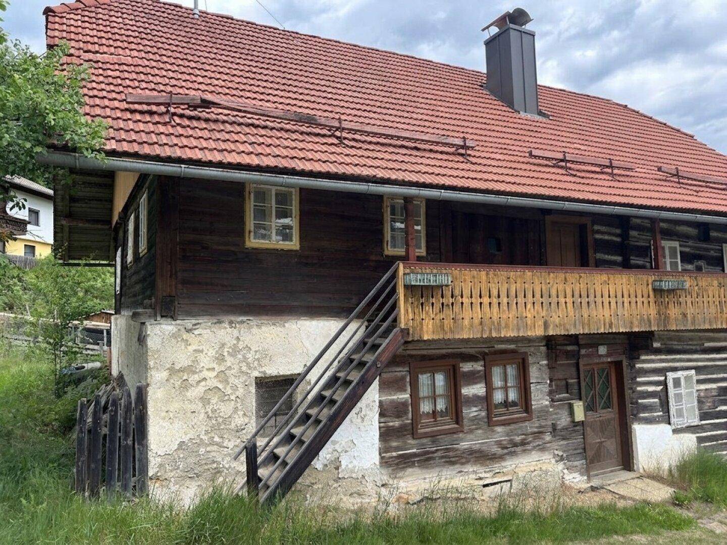 Rustikales Holzhaus mit rotem Ziegeldach, Holzbalkon und Außentreppe, eingebettet in grüne Landschaft.