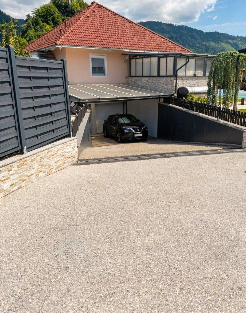 Einfahrt zum Haus mit Garage, Carport und rotem Ziegeldach in bergiger Landschaft.