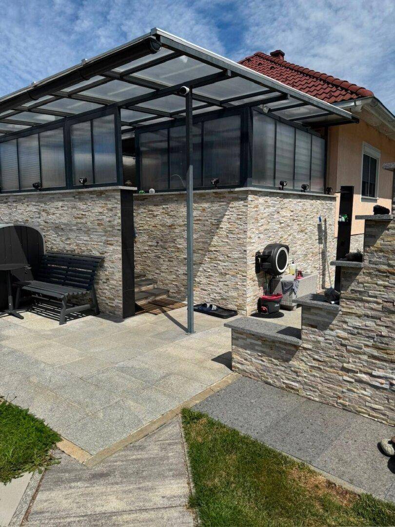 Überdachte Terrasse mit Steinfassade und Glasdach, angrenzend an das Wohngebäude.