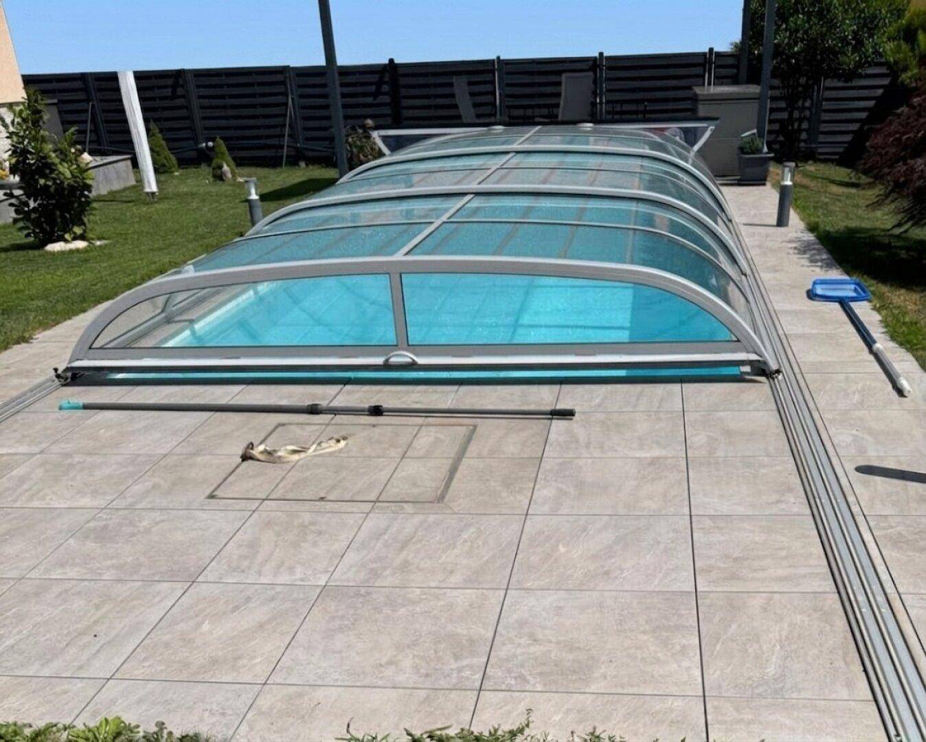 Überdachter Swimmingpool auf einer gefliesten Terrasse, umgeben von Rasenflächen.