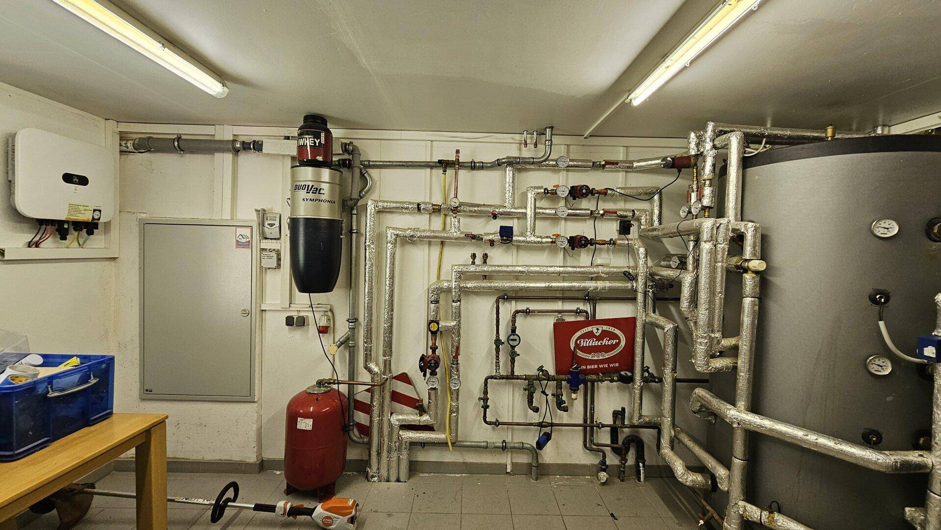 Weiterer Blick in den Technikraum mit zahlreichen Rohrleitungen, elektrischen Installationen und Lagerfläche.