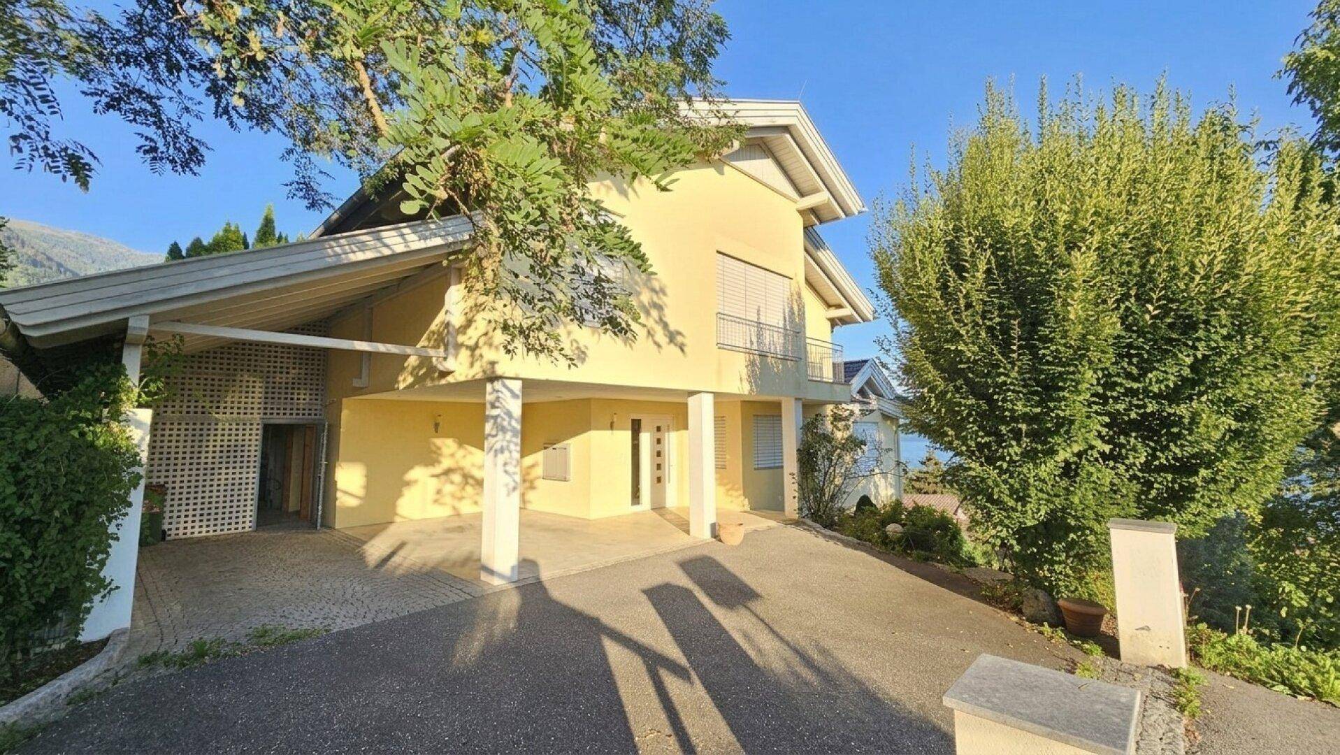 Zufahrt zum Haus mit überdachtem Carport und Eingangsbereich, umgeben von Bäumen und Sträuchern.