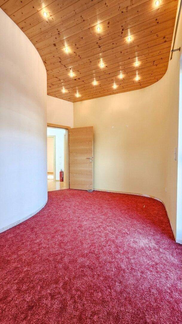 Zimmer mit auffälligem roten Teppichboden, geschwungener Wand und einer Holzdecke mit Einbaustrahlern.