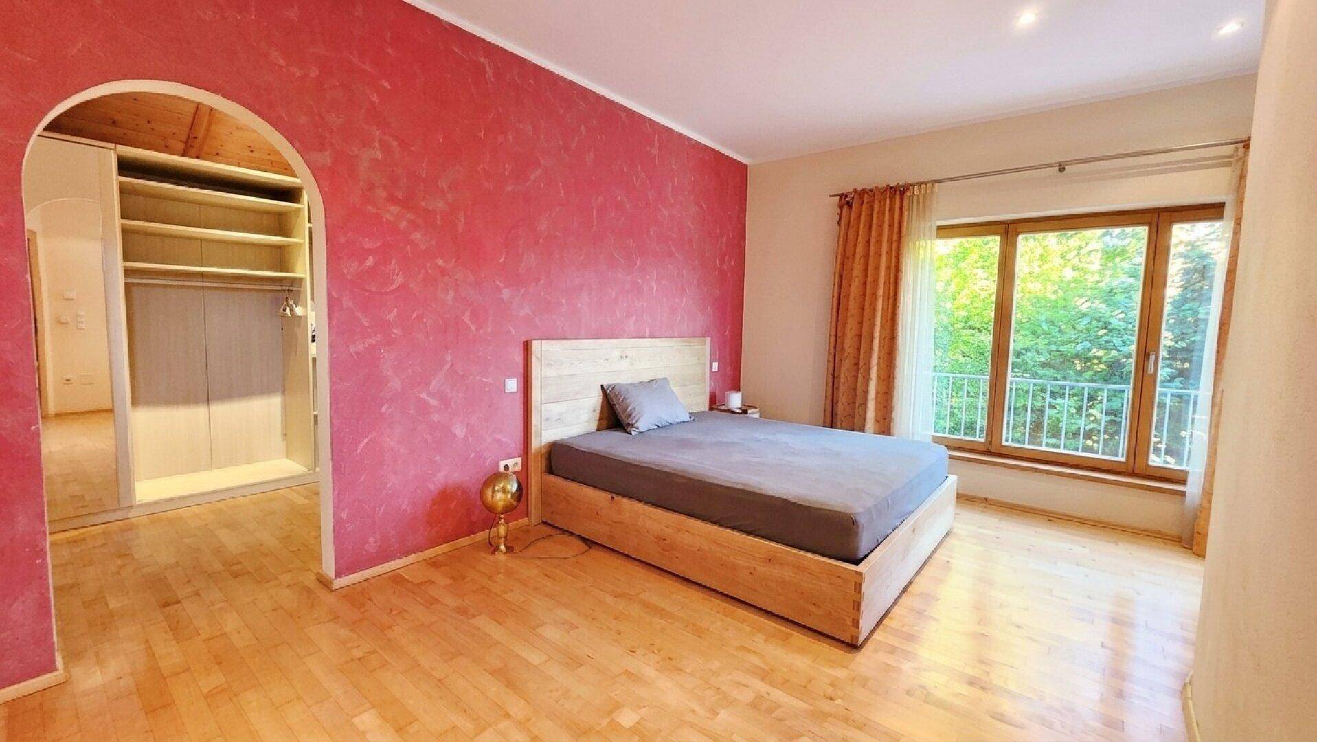 Gemütliches Schlafzimmer mit roter Akzentwand, Holzboden und Zugang zu einem französischen Balkon.