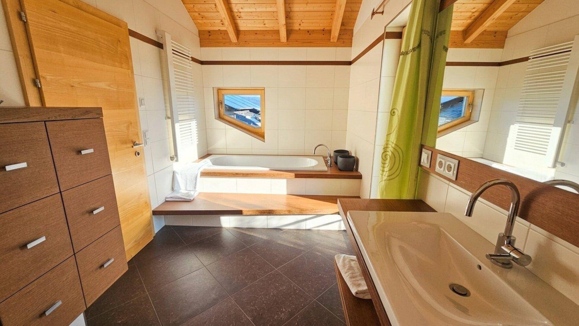 Geräumiges Badezimmer mit eingebauter Badewanne, Doppelwaschbecken und Fenstern unter der Holzdecke.