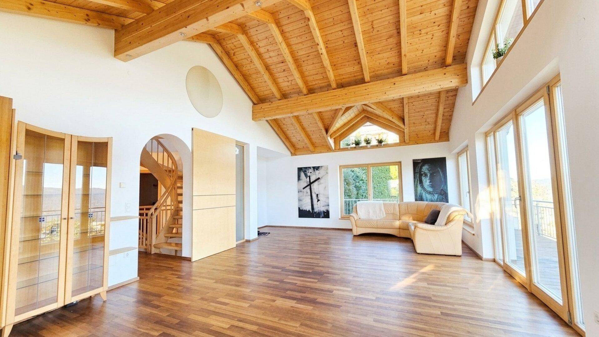 Helles Wohnzimmer mit beeindruckender Holzdecke, großen Fenstern und Zugang zu einer eleganten Holztreppe.