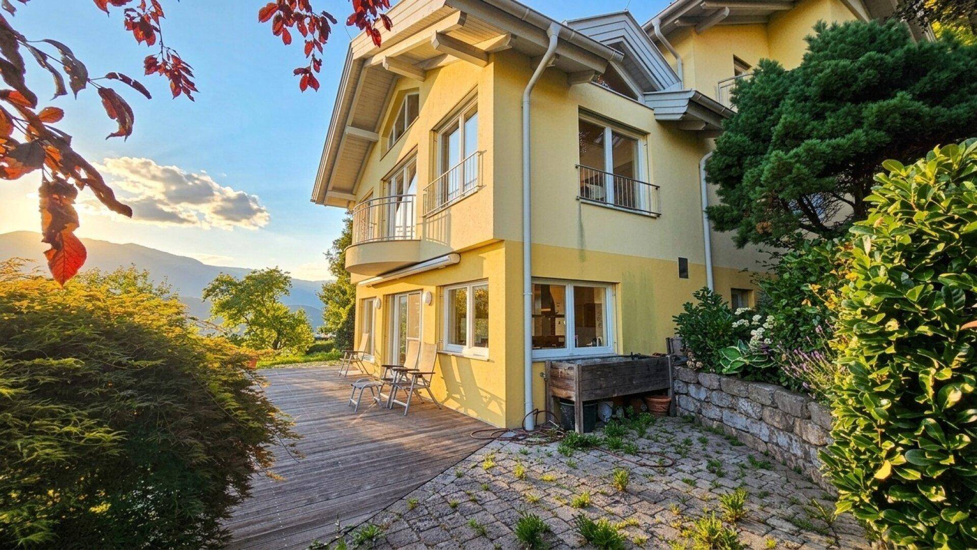 Charmantes gelbes Haus mit Holzterrasse, umgeben von üppigem Grün im warmen Licht der Abendsonne.