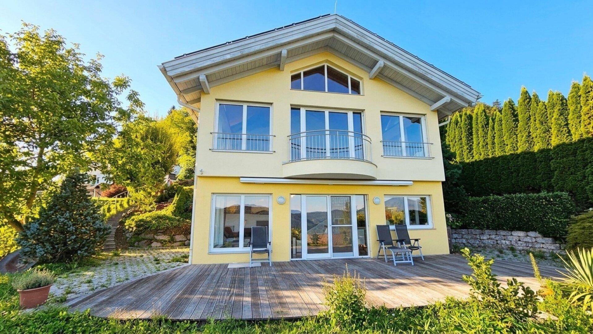 Frontansicht des modernen gelben Hauses mit weitläufiger Holzterrasse und gepflegtem Rasenbereich.