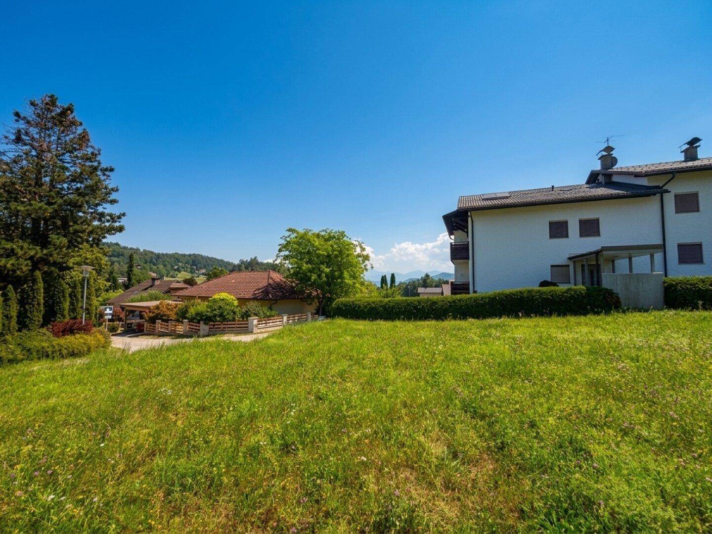 Offenes grünes Grundstück mit klarem Himmel, umgeben von Bäumen und Wohnhäusern in ländlicher Umgebung.