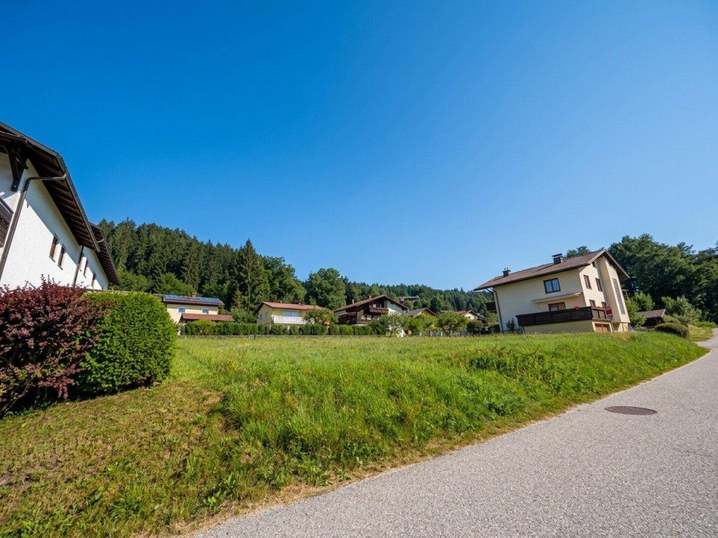 Unbebautes Grundstück am Straßenrand mit Blick auf bewaldete Hügel und benachbarte Wohnhäuser.