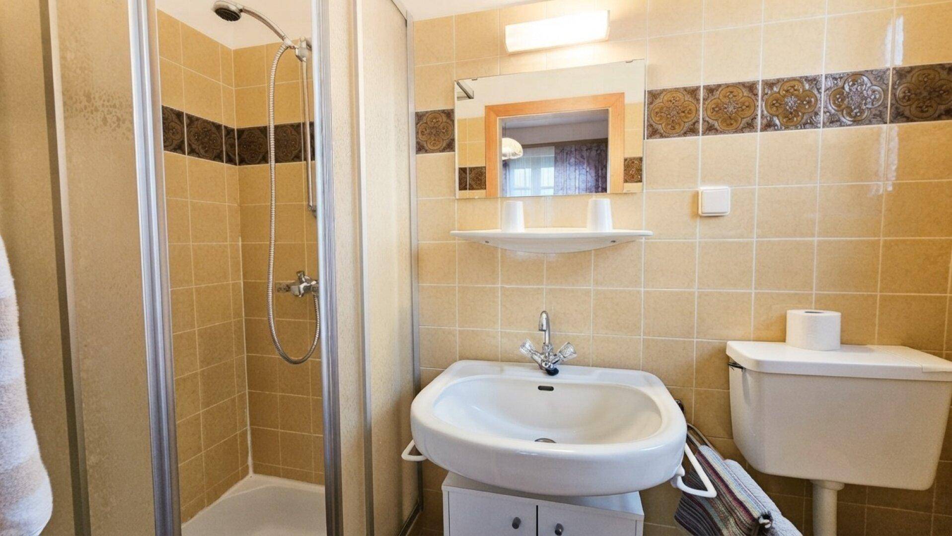 Badezimmer mit beigefarbenen Fliesen, Duschkabine und einem Waschbecken mit Spiegel.