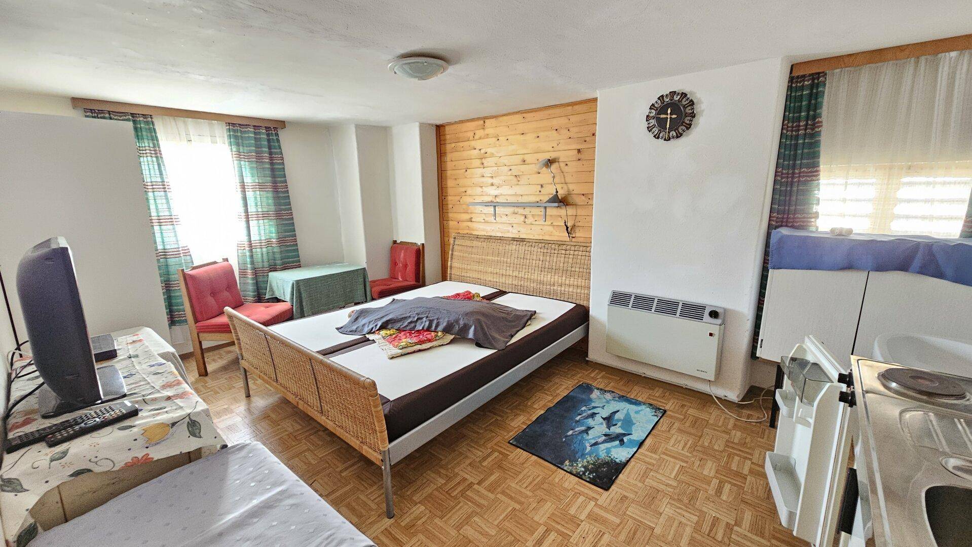 Gemütliches Schlafzimmer mit Doppelbett, Holzwand, kleiner Kochnische und karierten Vorhängen.