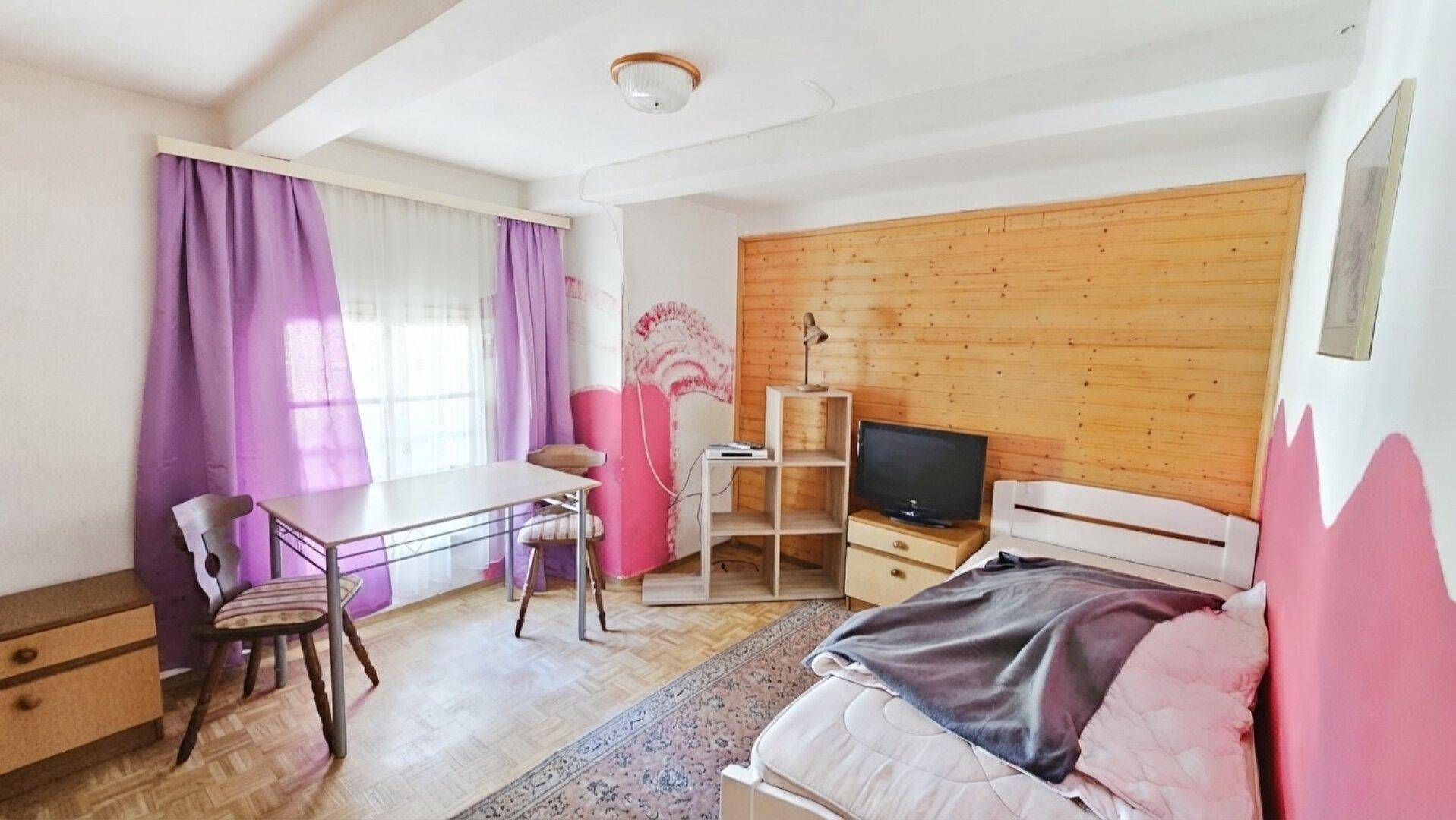 Schlichtes Schlafzimmer mit Holzwand, Einzelbett, Tisch und farbigen Vorhängen für eine gemütliche Atmosphäre.