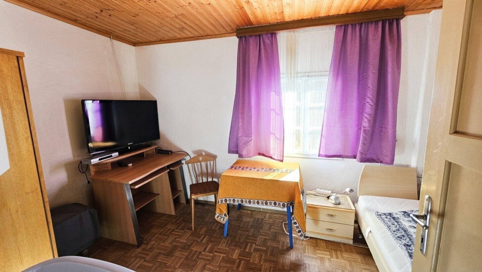 Einfaches Schlafzimmer mit Einzelbett, Schreibtisch, Holzdecke und lila Vorhängen für eine wohnliche Umgebung.