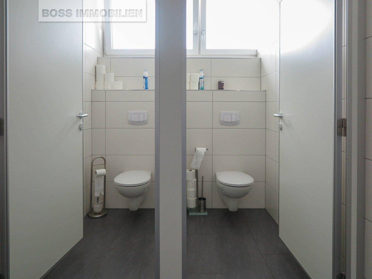 Zwei separate Toilettenkabinen in einem modernen Waschraum mit weißen Fliesen und dunklem Boden.