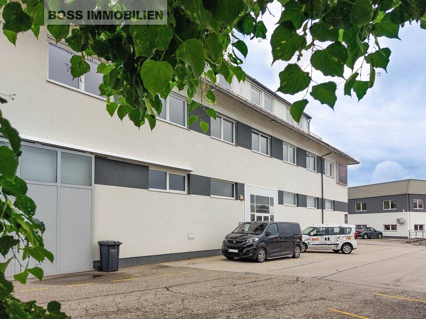 Außenansicht eines modernen Geschäftsgebäudes mit Parkplätzen und großen Fenstern, umgeben von Grün.