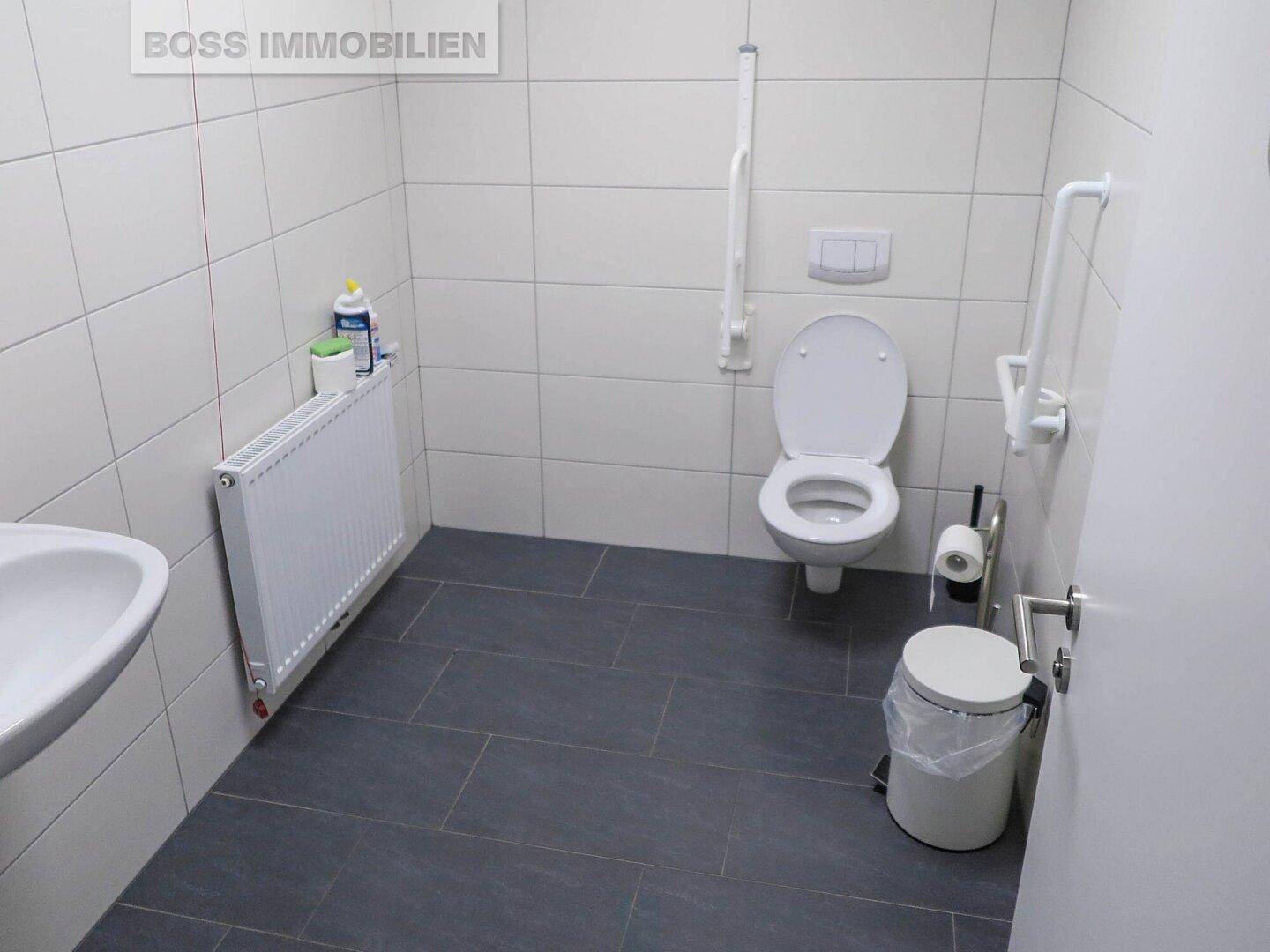 Barrierefreie Toilette mit Haltegriffen, wandhängendem WC, Waschbecken und Heizkörper in einem gefliesten Raum.