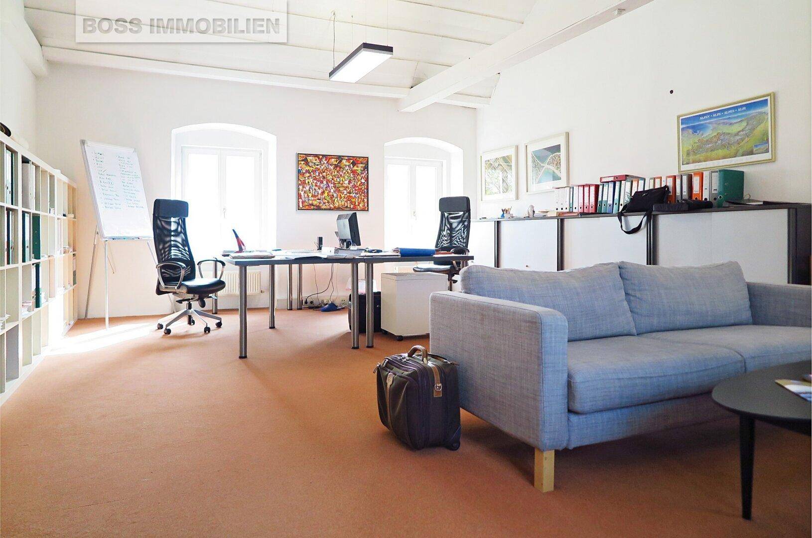 Büroraum mit Sofa, Schreibtischen, Bürostühlen und Regalen, auf einem orangefarbenen Teppichboden.