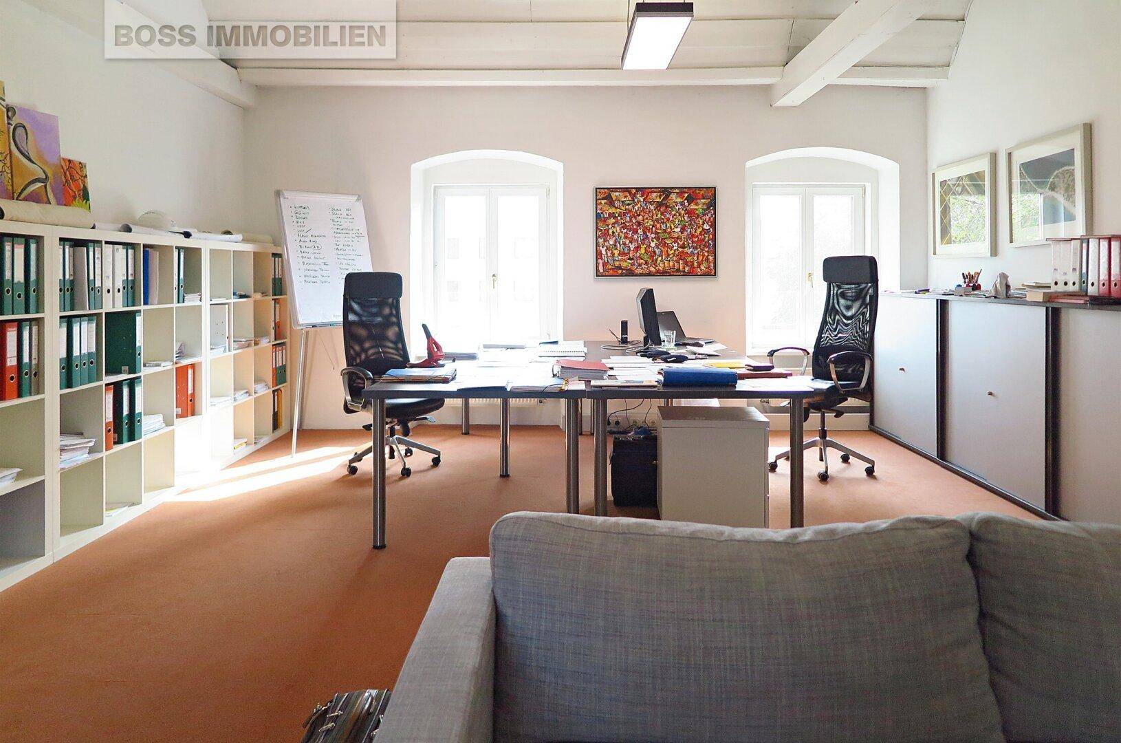 Büroraum mit Sofa, Schreibtischen, Bürostühlen und Regalen, auf einem orangefarbenen Teppichboden.
