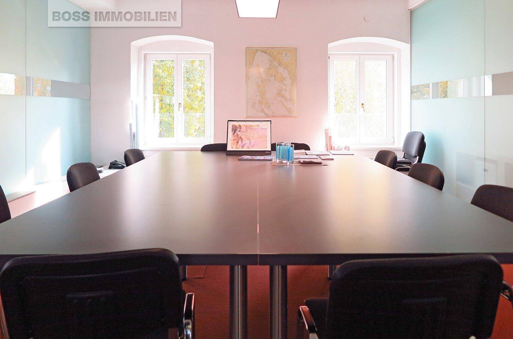 Konferenzraum mit großem Tisch, Stühlen und Fenstern, auf einem orangefarbenen Teppichboden.