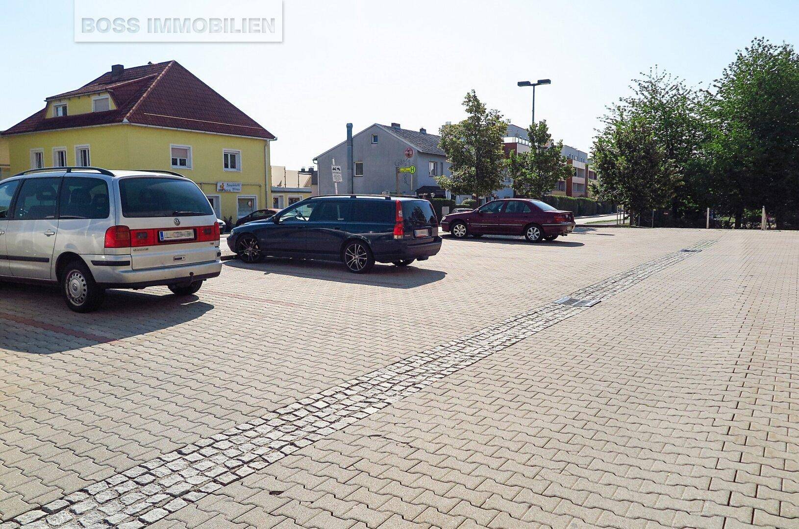 Großer gepflasterter Parkplatz mit mehreren Autos und angrenzenden Gebäuden unter blauem Himmel.