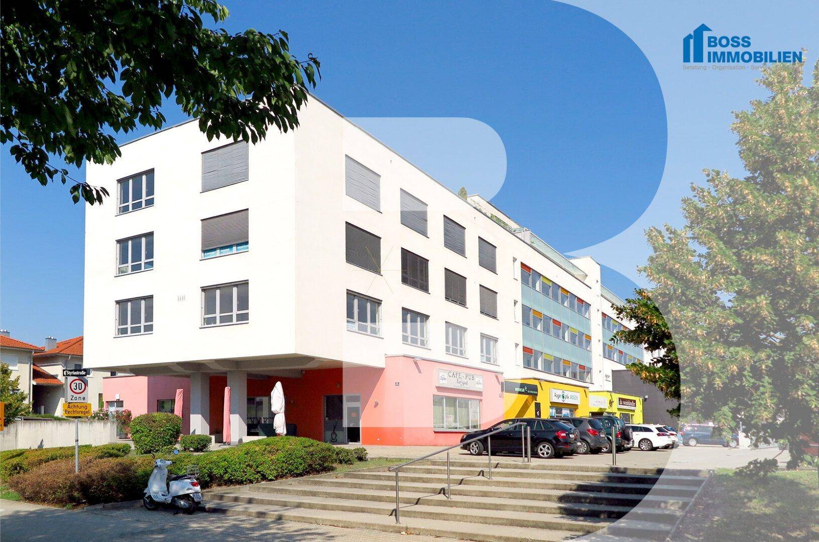Modernes Geschäftsgebäude mit farbigen Akzenten und Parkplätzen im Vordergrund, umgeben von Bäumen.