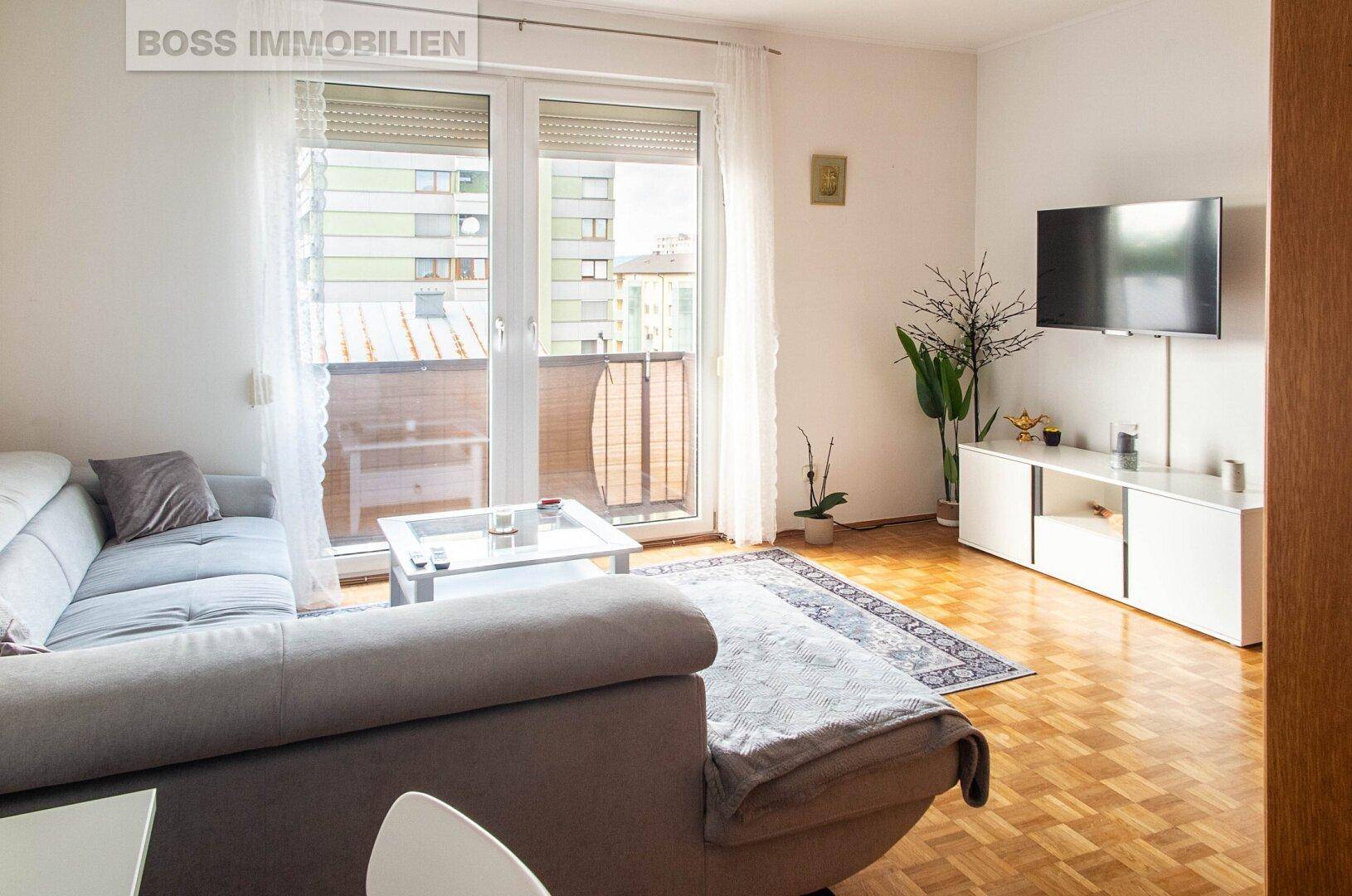 Helles Wohnzimmer mit Parkettboden, bequemer Couch und Zugang zum Balkon.