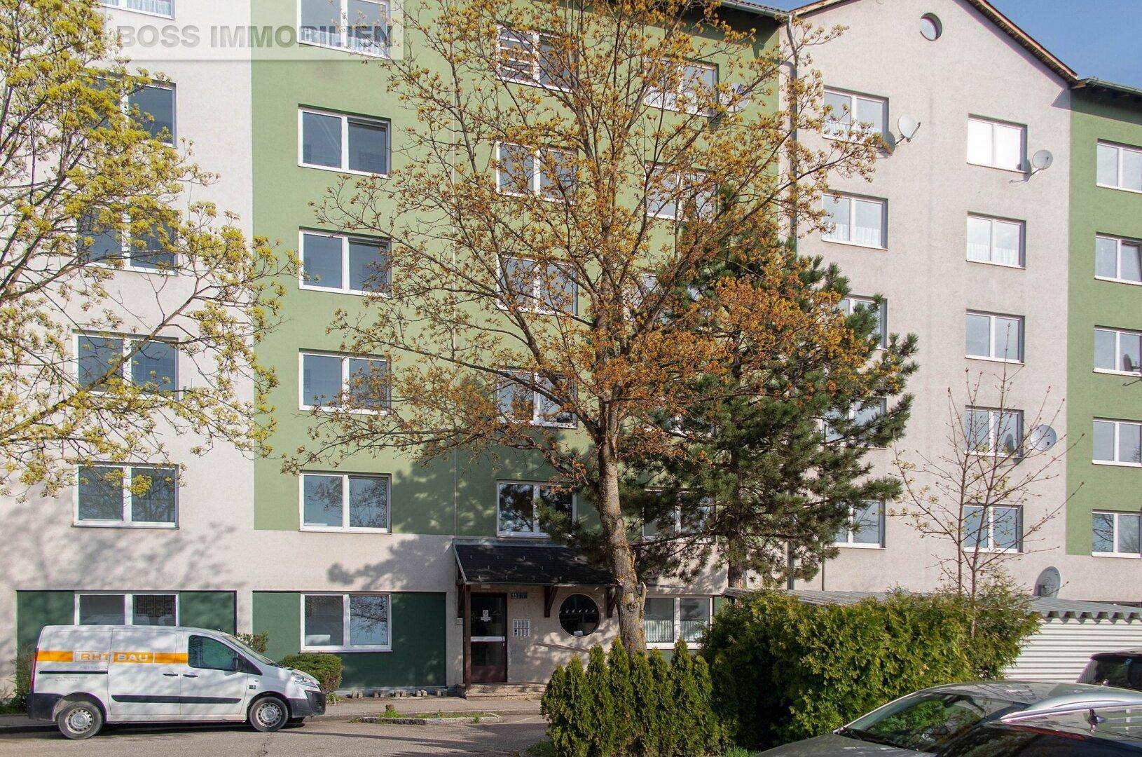 Mehrfamilienhaus mit grüner und beiger Fassade, umgeben von Bäumen und Sträuchern.