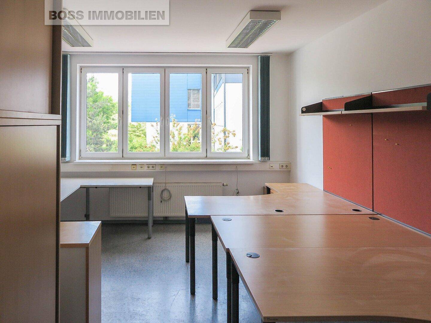 Funktionales Büro mit mehreren Arbeitsplätzen, großen Fenstern und einfacher Einrichtung, ideal für Teams.