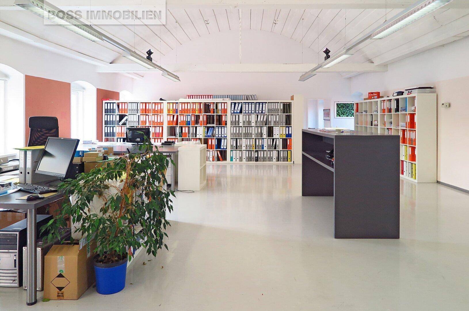 Weitläufiges Büro mit Schreibtischen, Bürostühlen und Regalen voller Aktenordner, unter einer hohen Decke.