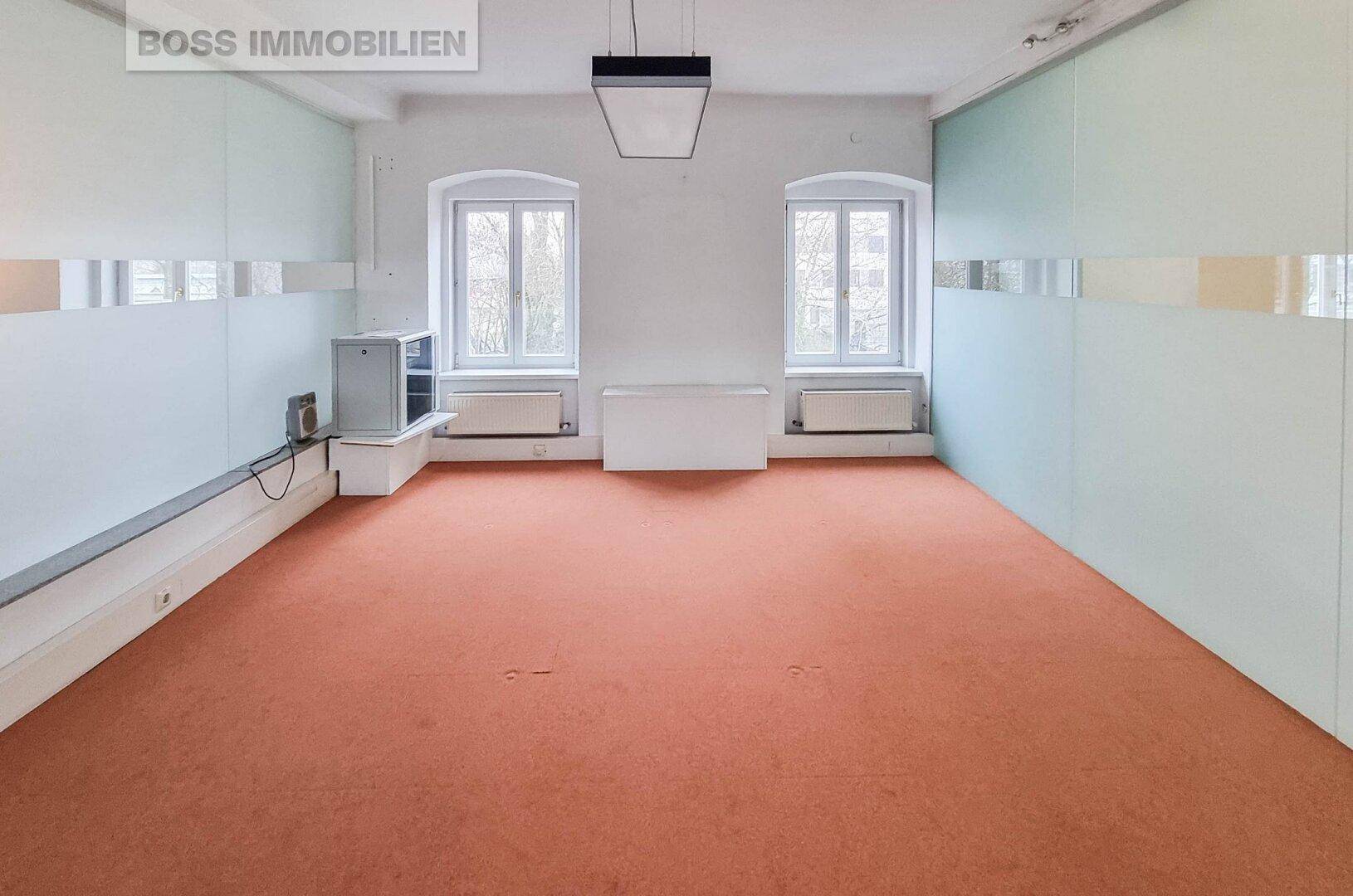 Leerer Büroraum mit zwei Fenstern, Heizkörpern, einem kleinen Kühlschrank und orangefarbenem Teppichboden.