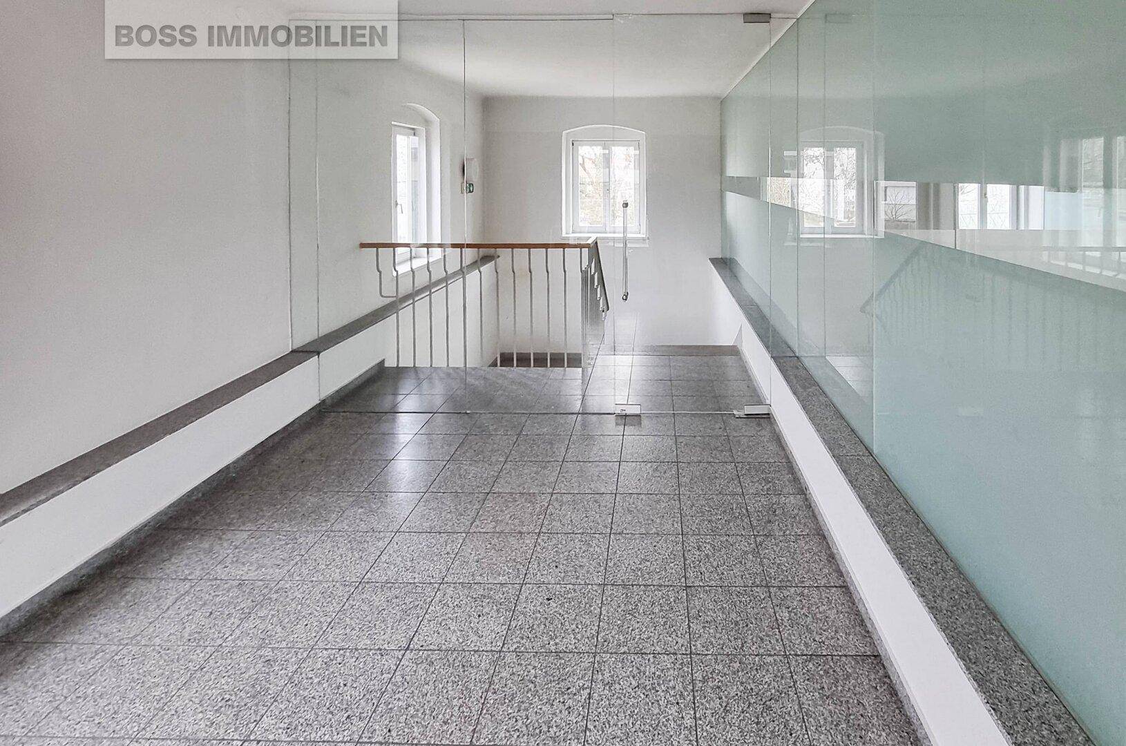 Heller Flur mit Terrazzoboden, Glasgeländer und Treppenaufgang zu weiteren Etagen.
