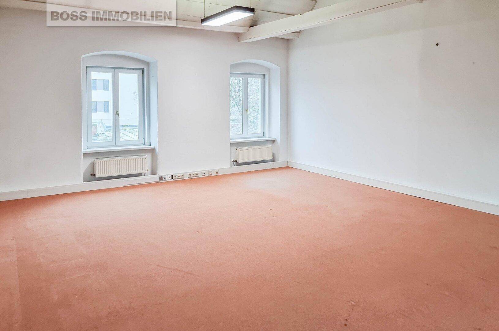 Leerer Büroraum mit zwei Fenstern, Heizkörpern und einem orangefarbenen Teppichboden.