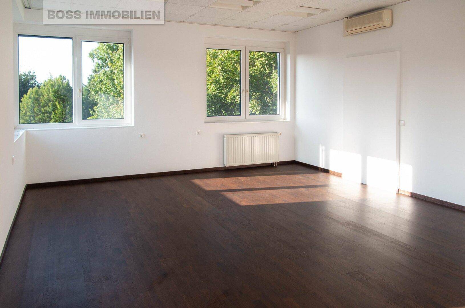 Geräumiger Büroraum mit dunklem Laminatboden, viel Tageslicht und Klimaanlage.