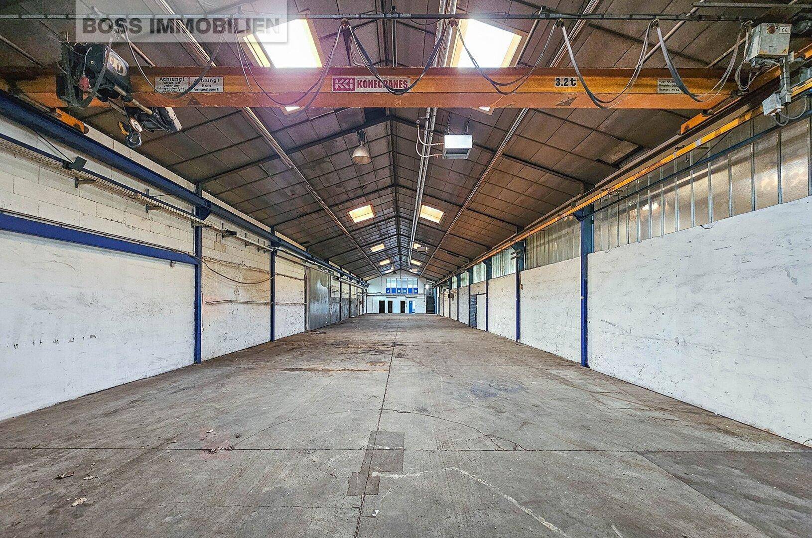 Große, leere Industriehalle mit Betonboden, hohen Decken und einem robusten Kran für schwere Lasten.