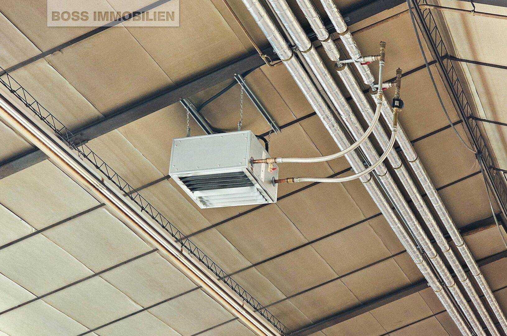 Deckenmontiertes Heiz- und Lüftungssystem mit sichtbaren Rohren in einem Industriebereich.